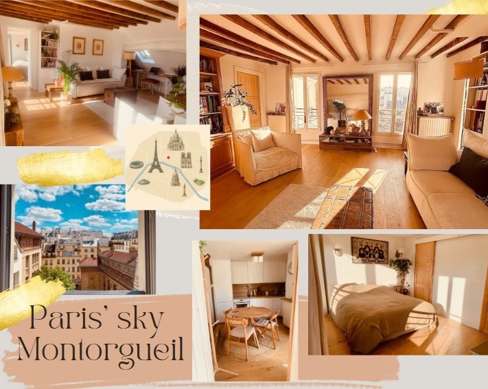 A sunlit living room showcases wooden beams and large windows that offer a view of the Paris sky. A cozy sofa sits beside a spacious armchair, with a dining area featuring a round table and chairs nearby. The kitchen is visible in the background.
