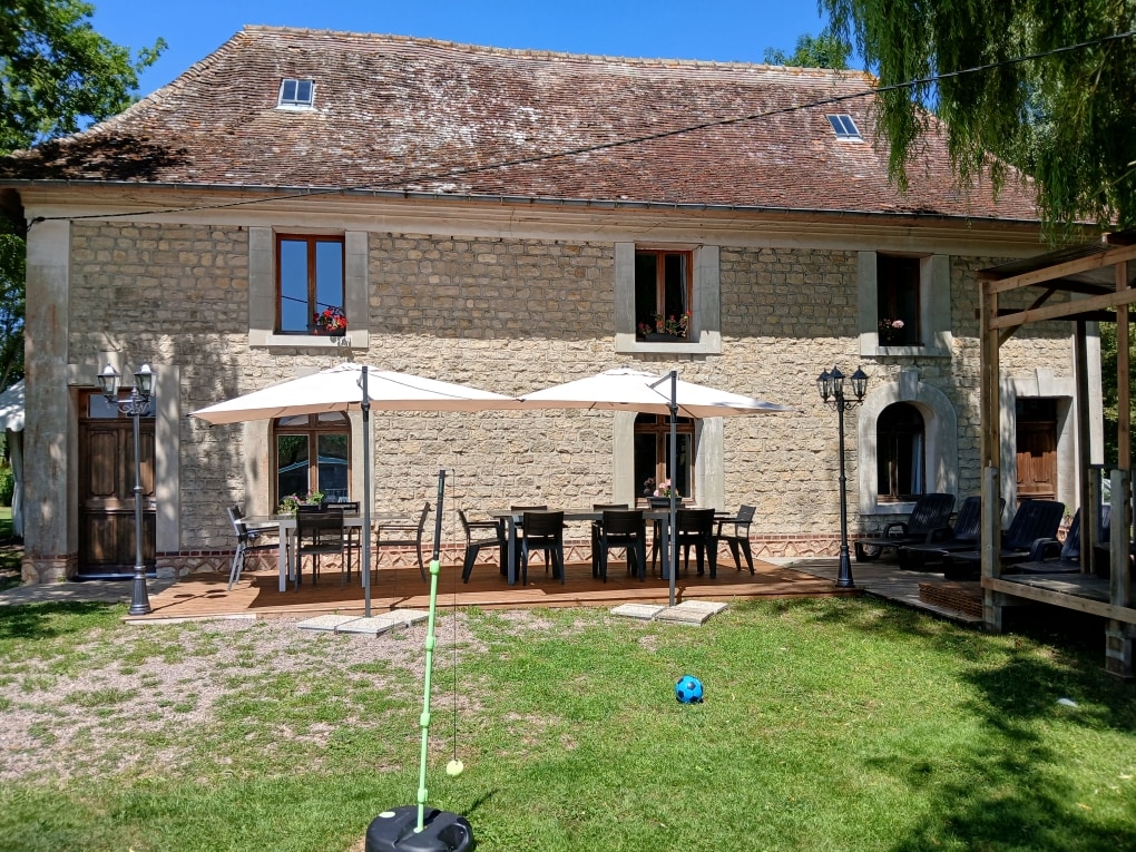 An outdoor dining area is positioned on a wooden deck, featuring a large table surrounded by chairs. Two patio umbrellas offer shade, while lounge chairs are arranged nearby. The building showcases a classic stone facade, with windows adorned with flower boxes.