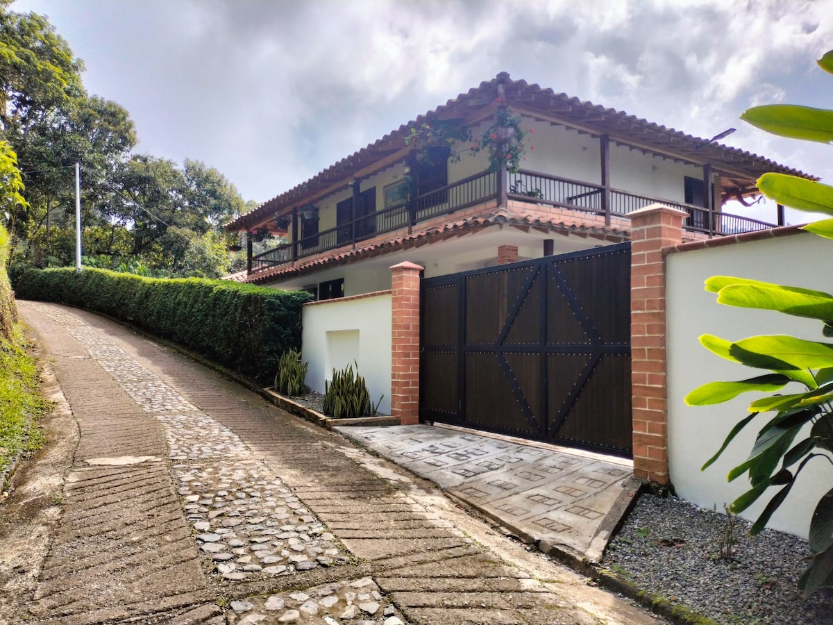 A charming two-story house is set amid greenery, with a sloped driveway leading up to a wooden gate. The structure features a traditional tile roof and is surrounded by lush plants, inviting guests to enjoy the tranquil outdoor setting.