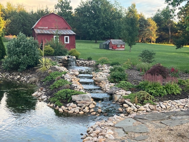 A serene landscape features a gently flowing waterfall leading to a pond, bordered by smooth stones. Lush greenery and colorful flower beds create a peaceful environment, with a classic red barn and a smaller structure visible in the background, enhancing the rural charm of the setting.
