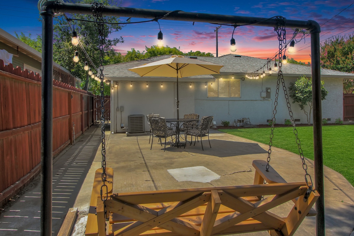 A cozy backyard features a wooden swing and a patio area with a table and chairs, all illuminated by string lights. An umbrella provides shade over the dining set, with a grassy space extending in the background. The home is visible in soft evening light.