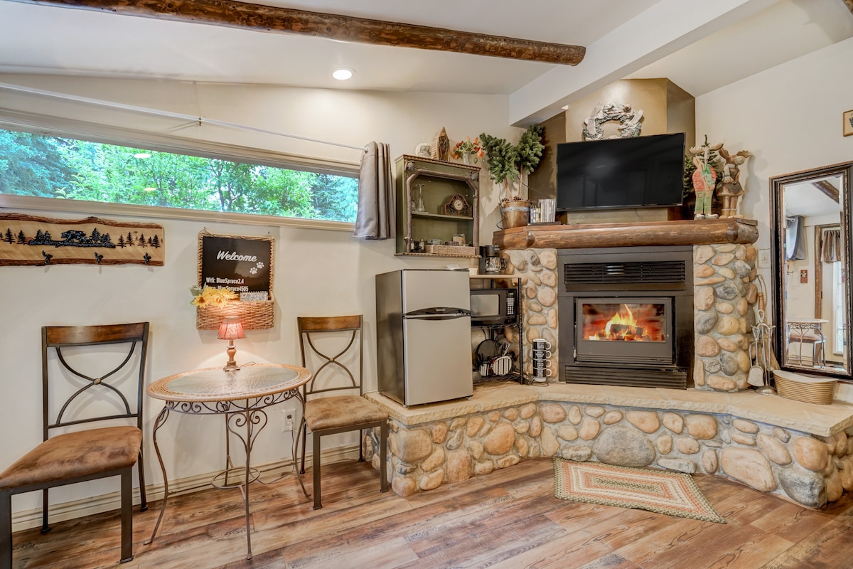 A welcoming living area features a stone fireplace with a small fire burning inside. A compact fridge is positioned beside a round table, which is accompanied by two chairs. Natural light enters through a large window, enhancing the cozy ambiance.
