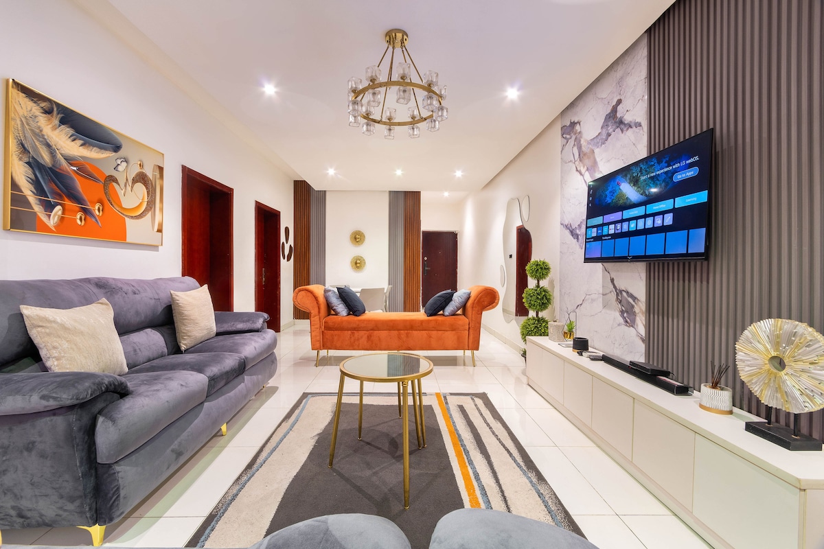A modern living area is presented, featuring a large gray sofa and an orange couch. A round coffee table is centered on a patterned rug, with a wall-mounted television displaying various options. Decorative plants and wall art complement the neutral-toned decor.