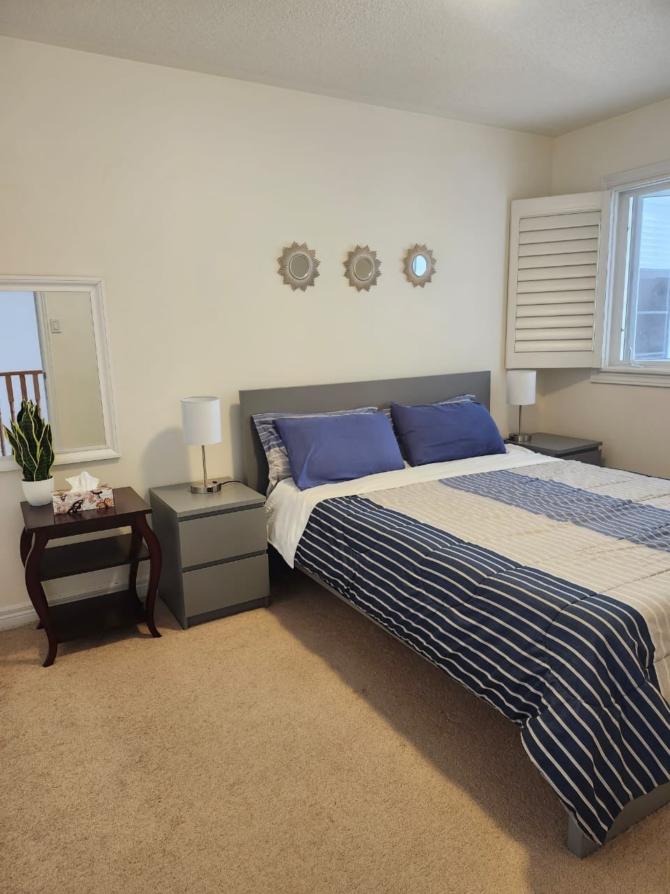 A private room featuring understated decor with a spacious bed dressed in white and blue bedding. Nightstands on either side hold lamps. A mirror and a small plant add decorative touches. Natural light enters through a window, complemented by wooden shutters.