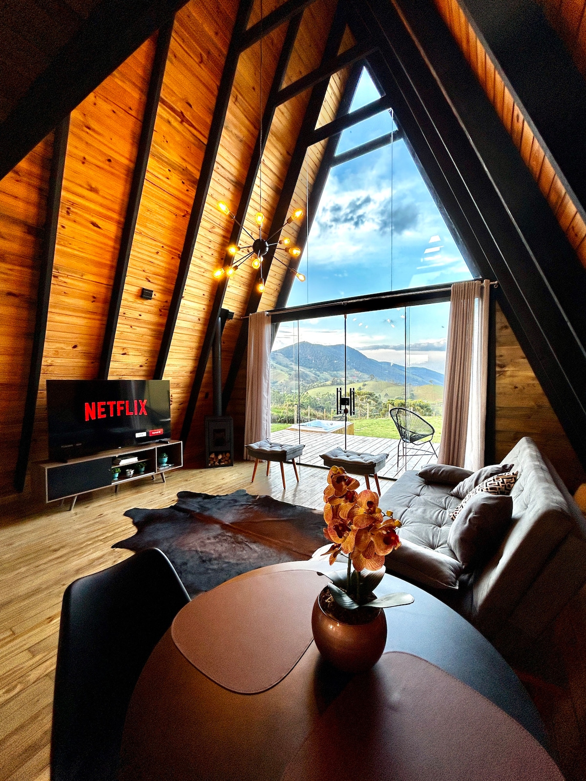 A spacious living area features a large window offering views of the valley and mountains. A dark leather sofa and a coffee table with a decorative vase are centered in the room. A television on a stand displays the Netflix logo, and natural wood accents enhance the design.