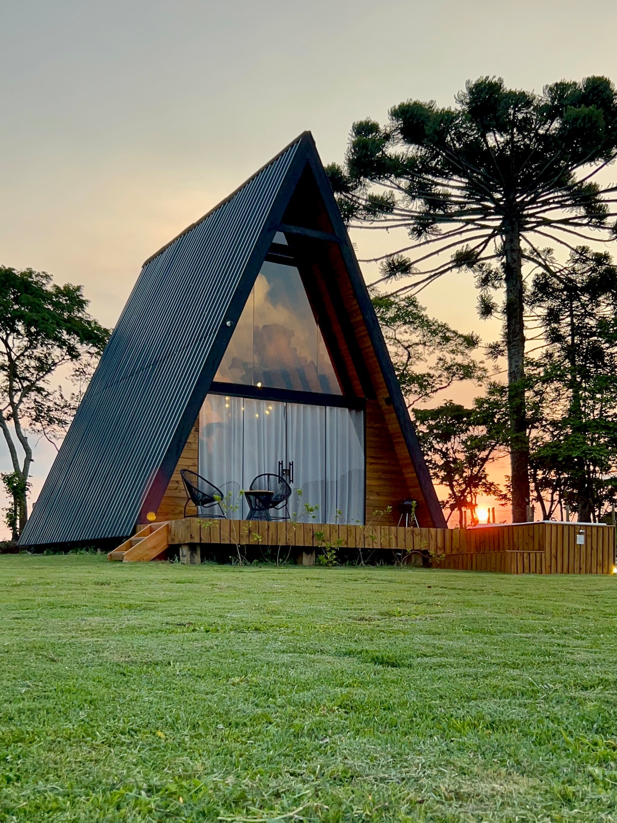The A-frame cabana is surrounded by green grass, highlighting its distinct triangular shape. Two outdoor chairs are positioned on the wooden deck, providing a cozy spot to enjoy the sunset. Tall trees complete the serene landscape, enhancing the natural ambiance.