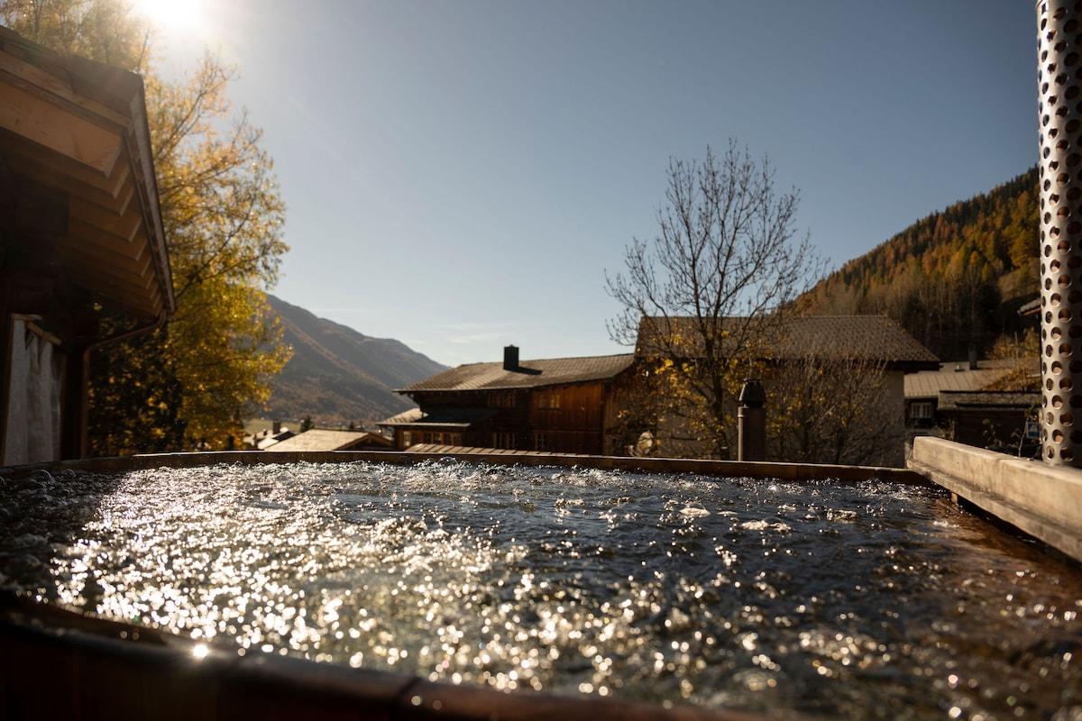 A hot tub is situated outdoors, surrounded by trees with golden leaves and a mountain backdrop. Sunlight reflects off the water's surface, creating a serene atmosphere. Nearby wooden structures complement the natural setting.