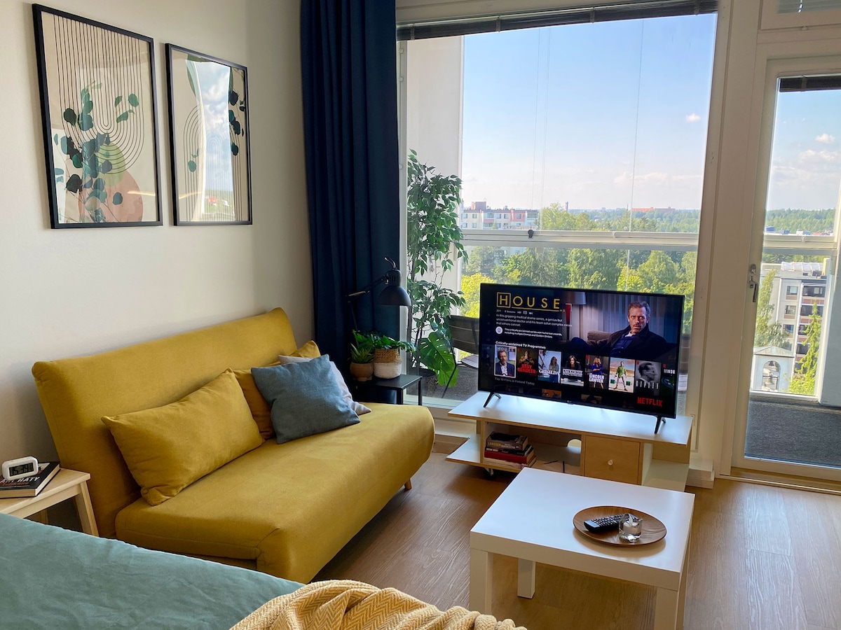 A cozy living area is presented, featuring a yellow sofa positioned next to a small white coffee table. A 43-inch smart TV displays content, while large windows provide ample natural light and a view of the surroundings. Framed artwork hangs on the wall beside the sofa.