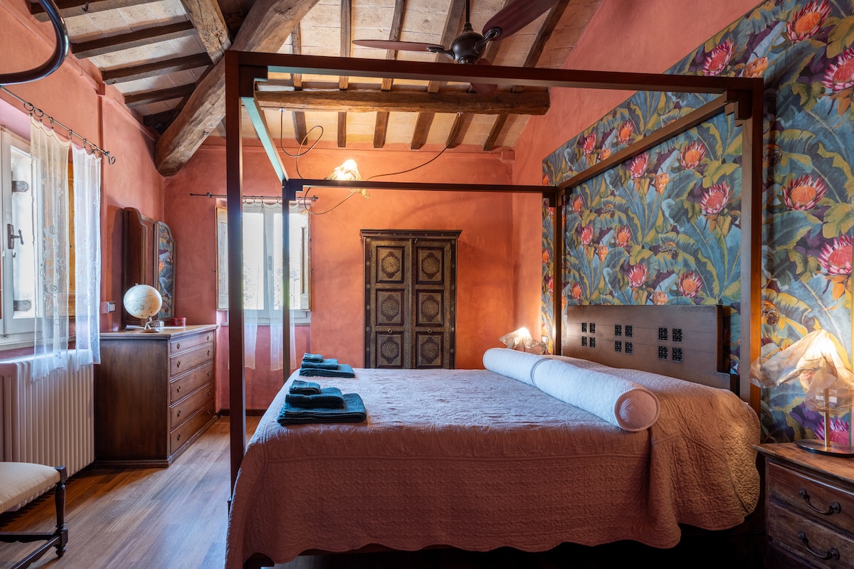 A tranquil bedroom features a spacious four-poster bed adorned with a light-hued coverlet. Decorative floral wallpaper sets the backdrop, complemented by wooden beams overhead. A wooden dresser and a round globe add character. Natural light filters through the window, enhancing the warm ambiance.
