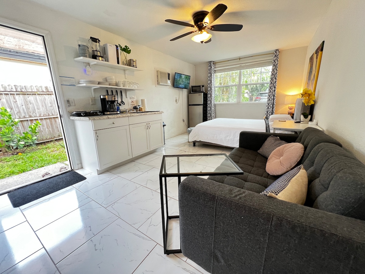 A modern studio space features a sleek kitchenette with white cabinetry and open shelving. A cozy couch is positioned next to a glass coffee table, while a spacious bed rests against the wall. Natural light enters through large windows, creating a bright and airy atmosphere.