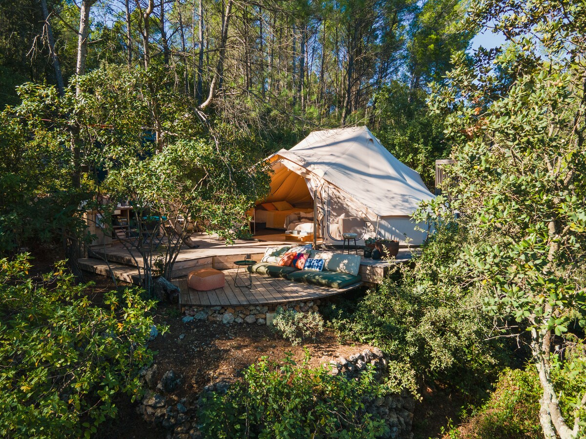 A spacious tent is positioned on a wooden terrace surrounded by greenery. Natural materials create a harmonious blend with the forest, while comfortable furnishings are visible. The entrance features large doors that can be opened, inviting the outdoors in.
