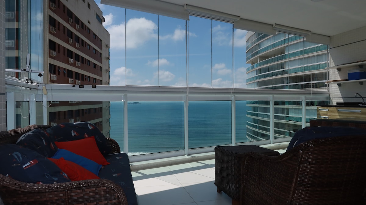 A balcony is shown with panoramic ocean views, featuring comfortable seating arranged around a table. Soft cushions in vibrant colors are placed on the seating. Large glass panels allow natural light to fill the space while providing an unobstructed view of the water and sky.