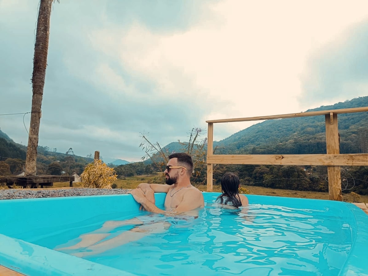 A spacious outdoor pool is surrounded by lush greenery and rolling hills. Soft clouds hang in the sky, and wooden railings frame the area, enhancing the serene atmosphere. Two individuals are enjoying the water, providing a sense of leisure and relaxation.