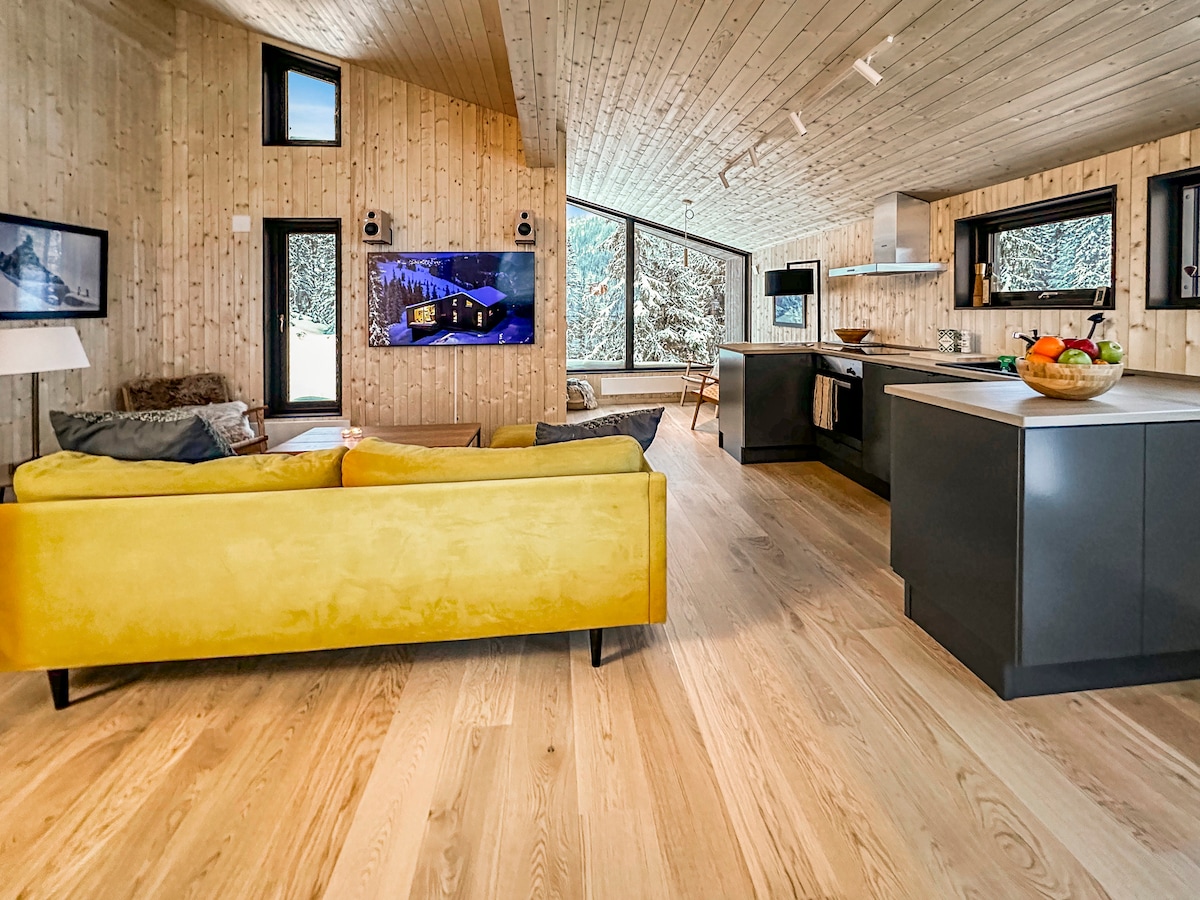An open living space is showcased, featuring wooden paneling and large windows that allow natural light to flood in. A yellow sofa offers a touch of color against the neutral tones, while a modern kitchen with sleek countertops and appliances is visible in the background.
