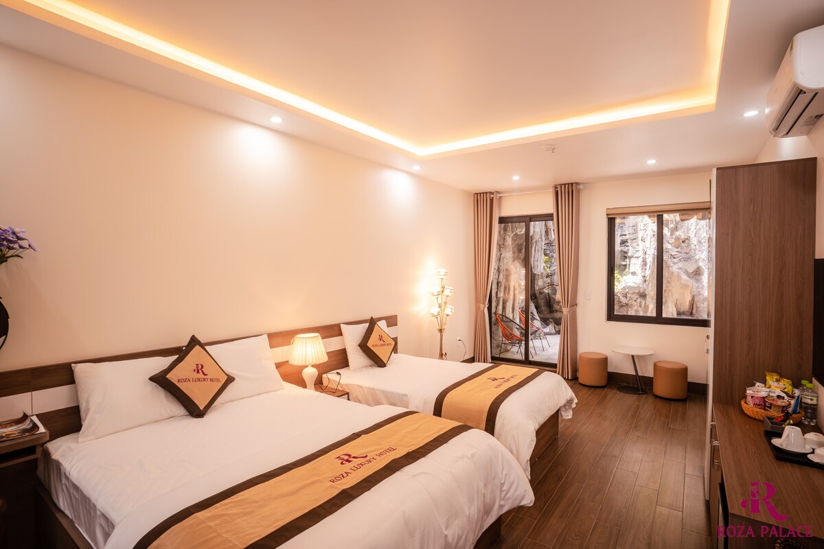 A spacious room is equipped with a double bed and a single bed, featuring modern decor. Natural light enters through large windows, while a small seating area is visible. A private bathroom can be accessed, and amenities are neatly arranged on a side table.