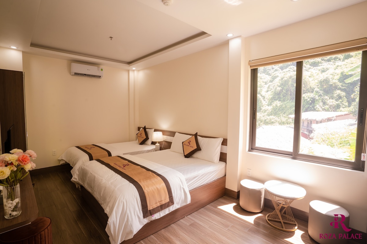 A spacious room features two neatly arranged beds with decorative throw pillows. Natural light fills the room through a large window, providing views of the outdoors. A dresser and seating area are visible, complemented by soft tones throughout the space.