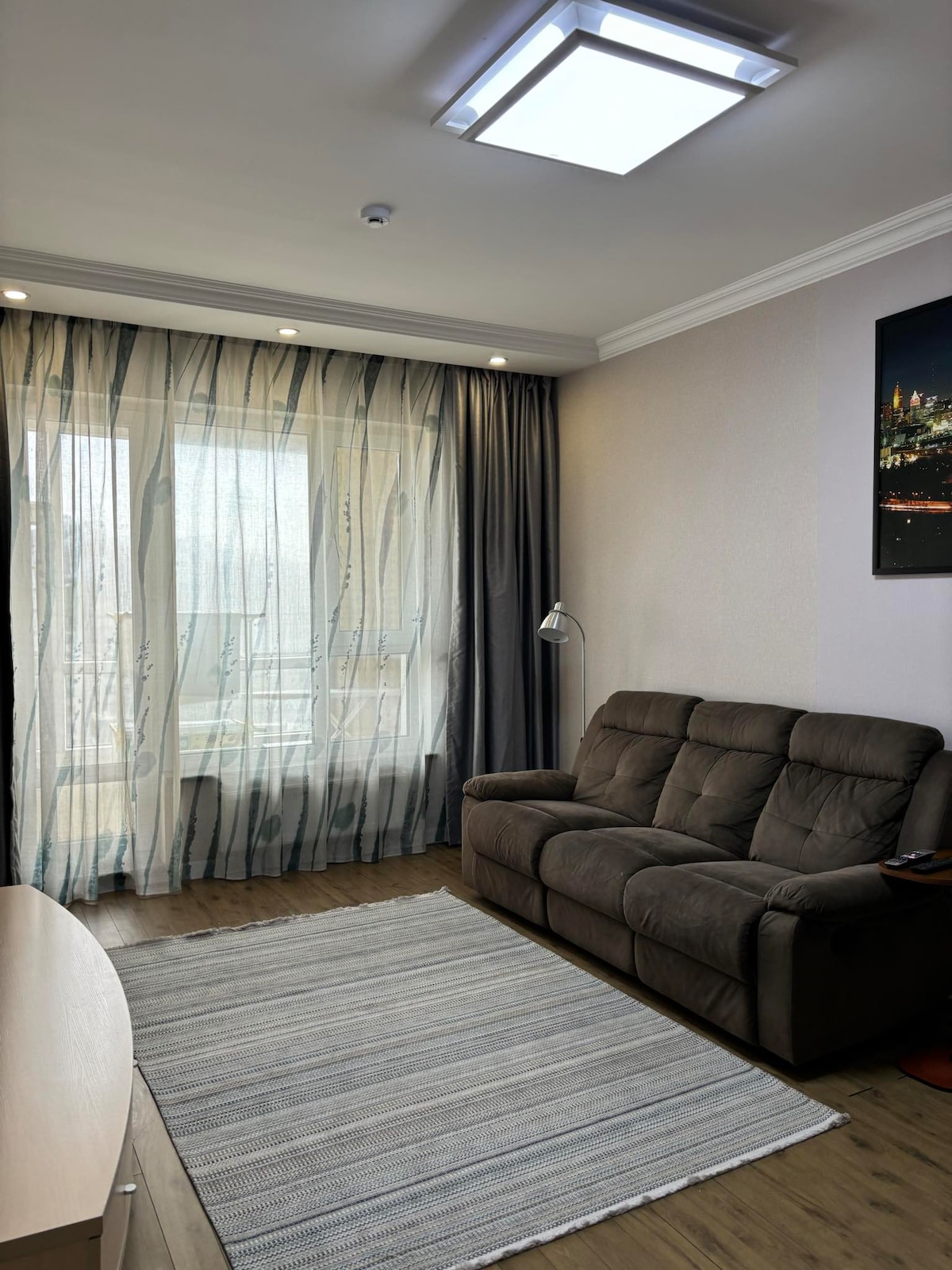 A living area features a comfortable brown sofa positioned beside a contemporary floor lamp. Sheer curtains drape over a large window, allowing soft light to fill the space. A light-colored area rug sits on the wooden floor, complementing the neutral wall tones.