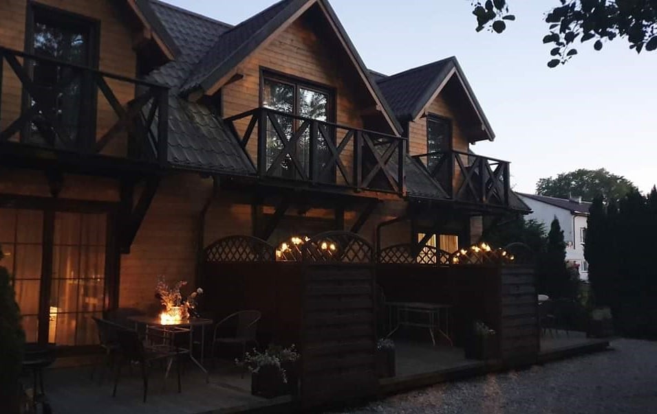 The exterior of the wooden cabins is shown, featuring a cozy arrangement of balconies and outdoor seating areas. Soft lights illuminate the front, creating a welcoming evening atmosphere. Lush greenery surrounds the space, enhancing the tranquil environment.