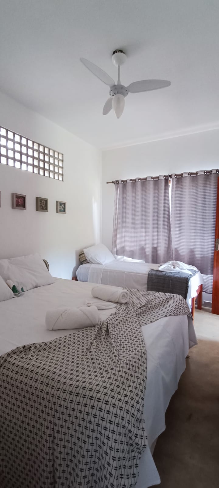 A bright sleeping area is highlighted by natural light filtering through sheer curtains. The room features a spacious double bed and a single bed, both dressed in crisp white linens. Textiles include a patterned throw and neatly rolled towels, contributing to a tidy appearance.