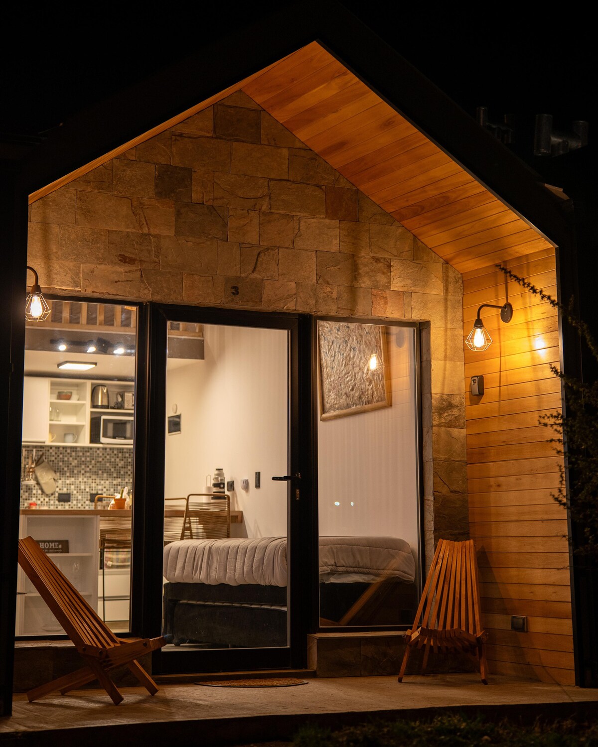 A charming entrance is illuminated by warm outdoor lighting. Glass doors reveal a cozy interior with a bed and kitchenette. Two wooden chairs are positioned on the porch, enhancing the inviting atmosphere of the space.