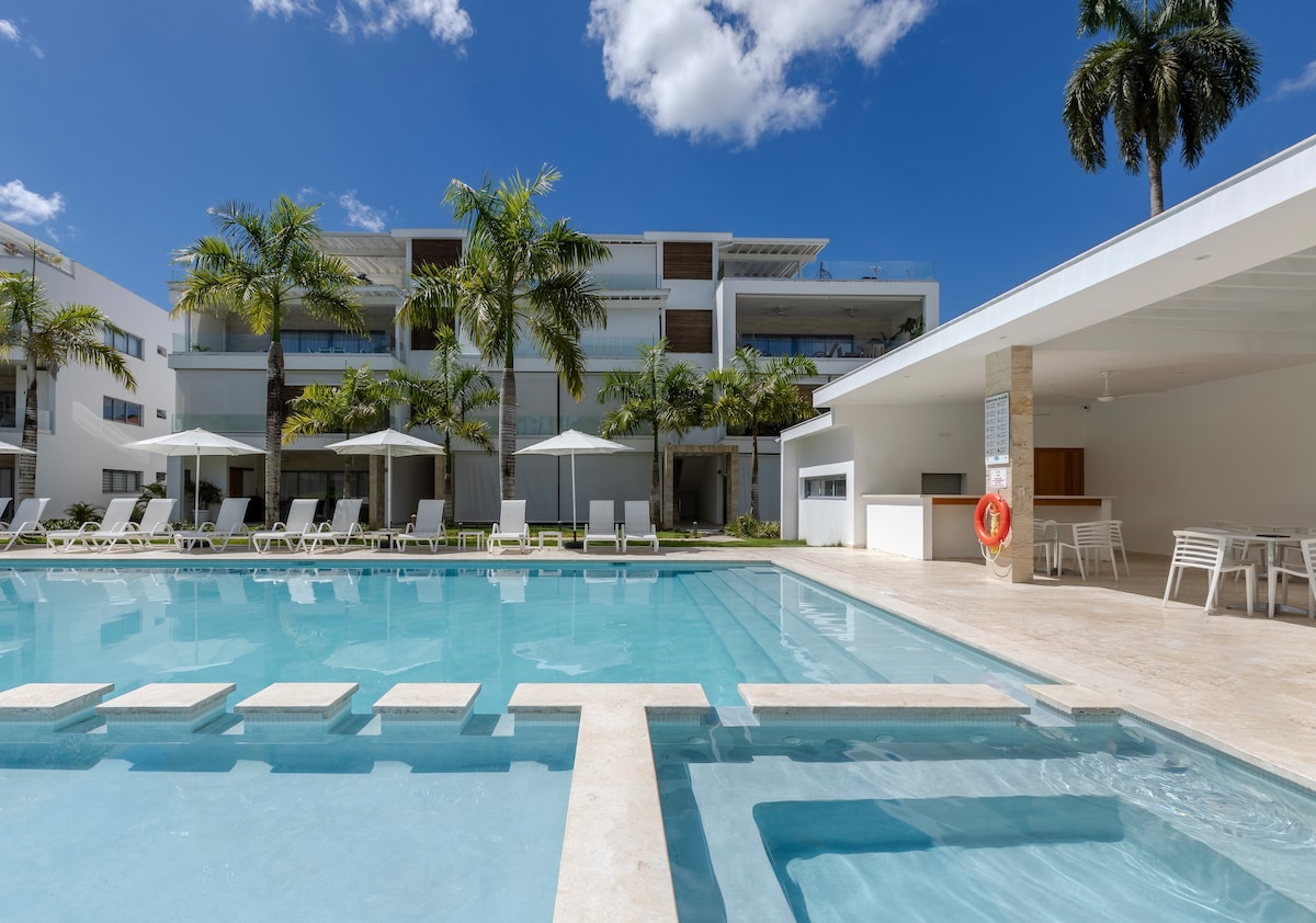 The image showcases a sparkling outdoor pool surrounded by palm trees. Several lounge chairs are positioned under large umbrellas, offering shade. The modern architecture of the building features large glass doors and balconies, creating a seamless flow between indoor and outdoor spaces. A barbecue area is visible to the right.