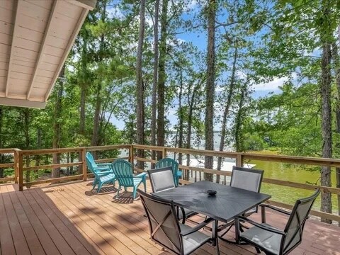 A spacious deck is furnished with a black table surrounded by six chairs, complemented by bright blue and green lounge chairs. A view of the serene water can be seen through the green trees, offering a peaceful outdoor space for relaxation.