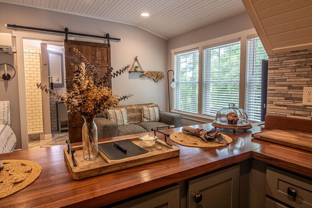 The open kitchen features warm wood surfaces with a breakfast bar, natural decorations, and a cozy seating area with a sofa. Large windows provide ample natural light, while a sliding barn door leads to the spa-inspired bath. A peaceful atmosphere is created with soft textures.