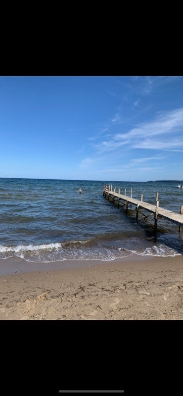 A wooden pier extends into calm waters under a clear blue sky, with gentle waves lapping at the sandy shore. A person is seen near the water's edge, enjoying the serene coastal environment.
