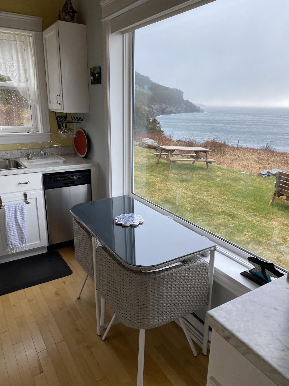 A kitchen area features a modern table with a glass top, positioned near a large window that provides a view of the North Atlantic Ocean. Outside, a picnic table and chairs are visible on a grassy area, framed by coastal hills under overcast skies.