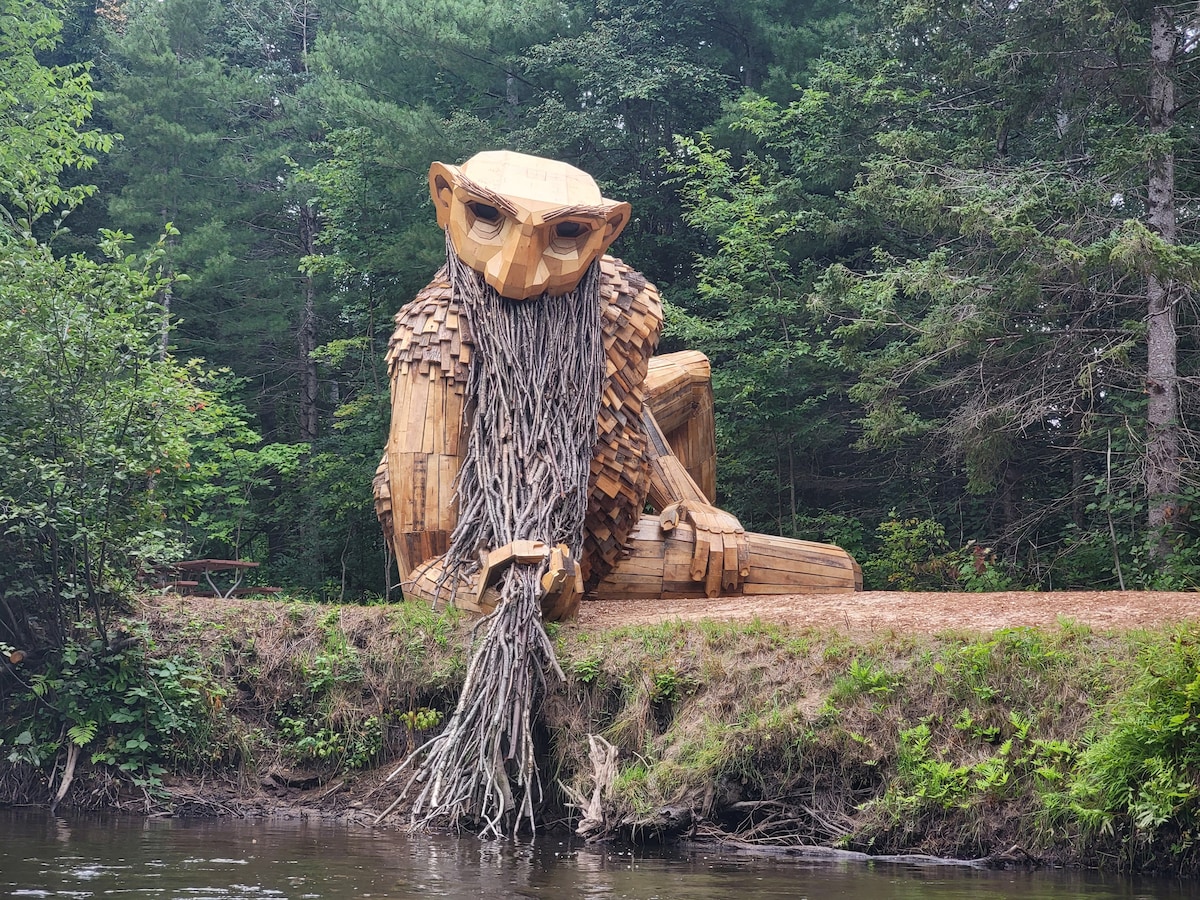 A large wooden sculpture resembling a lion sits at the edge of a lush forest, crafted from logs and branches. Its flowing mane is made of intertwined twigs, while it overlooks a serene riverbank surrounded by greenery.
