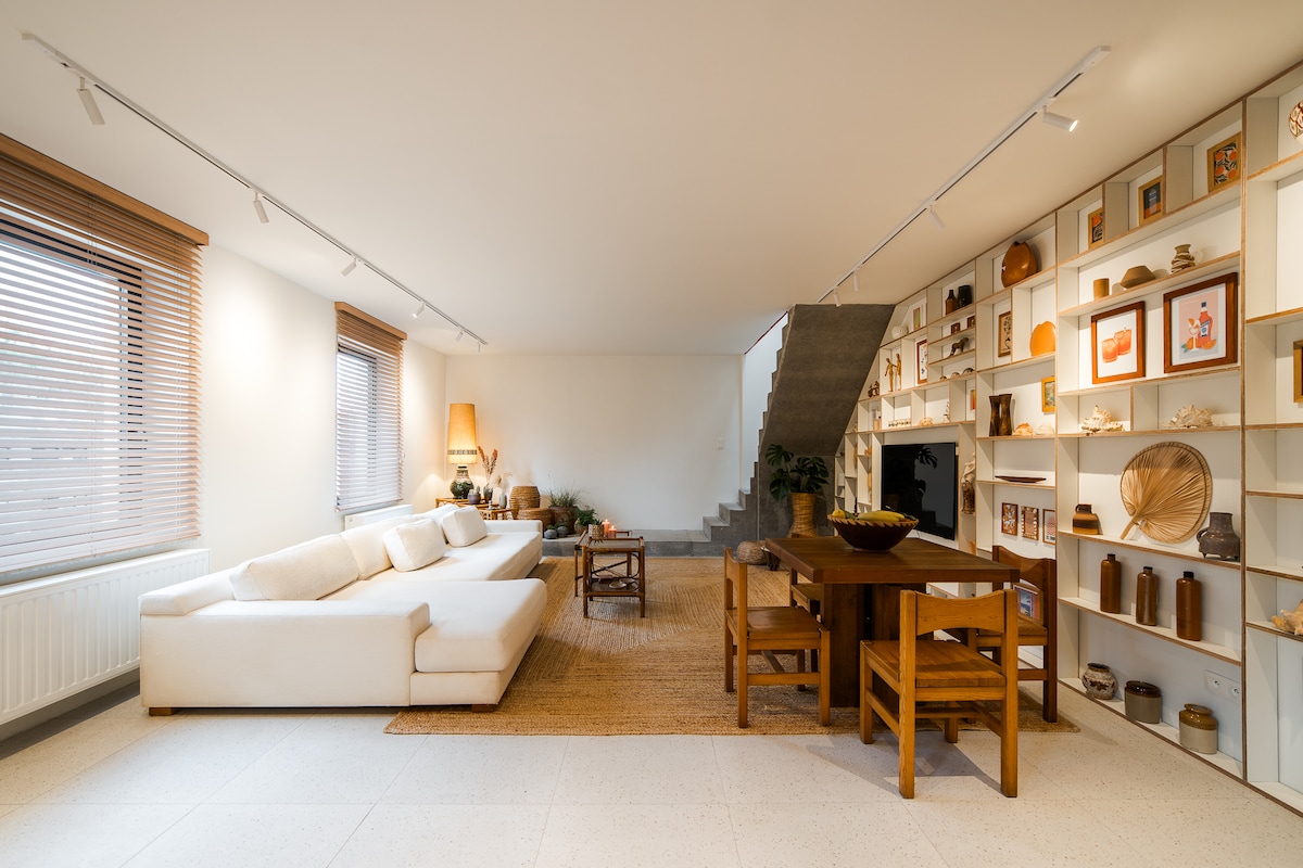 The spacious living area features a white sectional sofa paired with a wooden coffee table. A dining table with wooden chairs is positioned nearby. Natural light enters through the windows, while decorative shelves display an array of curated items. A staircase leads to the upper level.