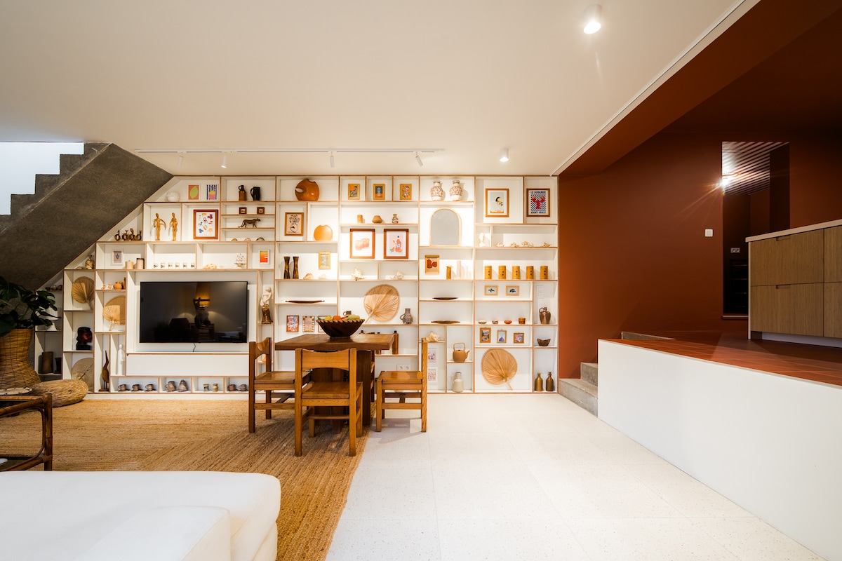 A living area features a warmly decorated wall with various art pieces and decorative items. A wooden dining table with matching chairs is positioned nearby, while a sleek television is mounted on the shelf. Soft lighting enhances the inviting atmosphere, complemented by a modern kitchen area in the background.