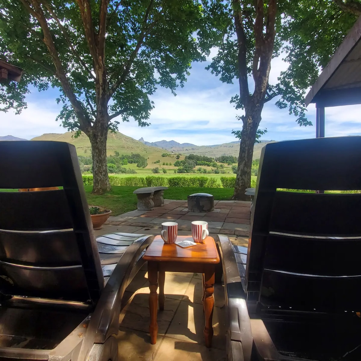 A pair of black lounge chairs are positioned facing a scenic view, framed by large trees. In front, a small wooden table holds two cups, inviting guests to enjoy the peaceful surroundings. The distant mountains and open fields create a serene backdrop.