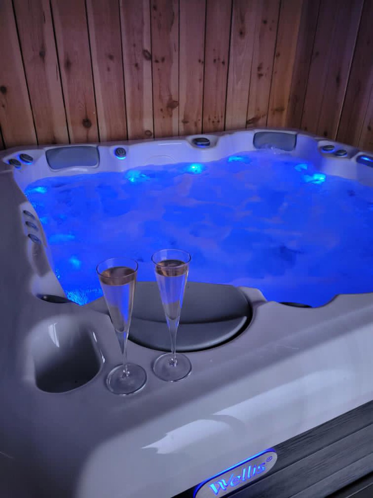 A well-appointed spa features a hot tub illuminated with soft blue lights. Two glasses are positioned on the edge, inviting relaxation. The wooden paneling complements the serene atmosphere, creating a soothing environment for unwinding.