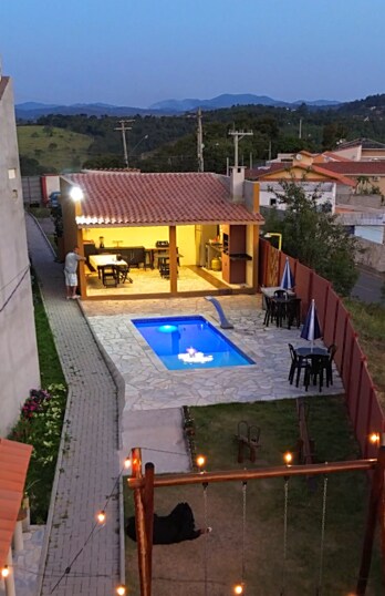 An elevated view of the outdoor area features a swimming pool surrounded by a stone patio. A cozy outdoor dining setup with tables and umbrellas is visible, along with a nearby grill area. Lush greenery and mountains form the backdrop, enhancing the serene atmosphere.