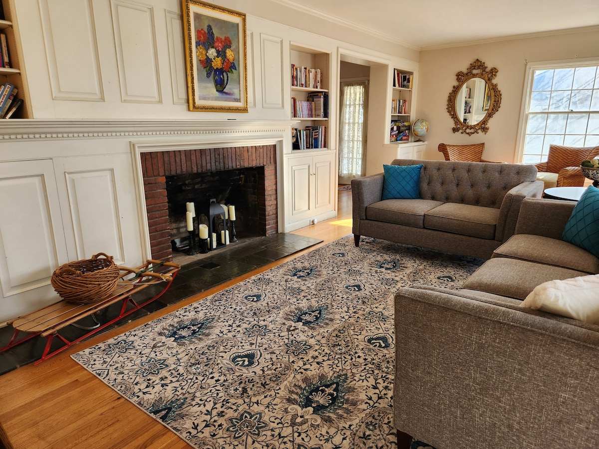 A welcoming living room features a stone fireplace with candles and decorated shelving. Two gray sofas are arranged around a central area rug that displays a floral pattern. Natural light streams through a large window, illuminating the space.