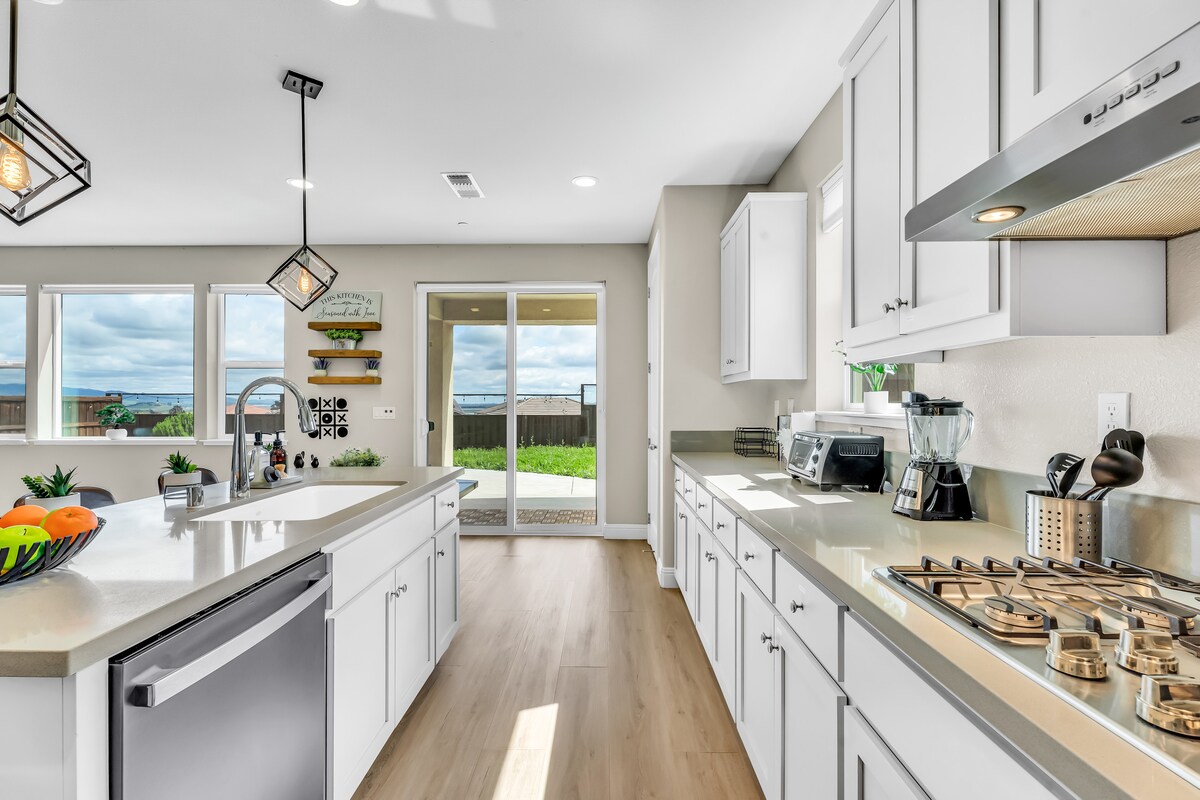 A modern kitchen features sleek white cabinetry, stainless steel appliances, and a spacious countertop. Natural light pours in through large windows, offering views of the outdoor area. A decorative bowl of fruit is arranged on the island, enhancing the room's inviting atmosphere.