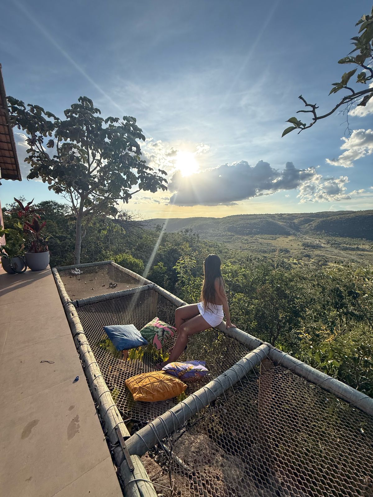 A tranquil outdoor space features netted seating overlooking a lush landscape. Sunlight brightens the scene as it descends behind distant hills, creating a serene ambiance. Decorative cushions in vibrant colors are positioned nearby, inviting relaxation amidst nature.