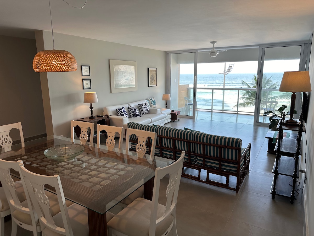 A spacious living area features large glass windows providing ocean views. A wooden dining table is accompanied by six white chairs. A comfortable seating arrangement includes a striped sofa and decorative pillows. Soft lighting is provided by hanging fixtures and table lamps.