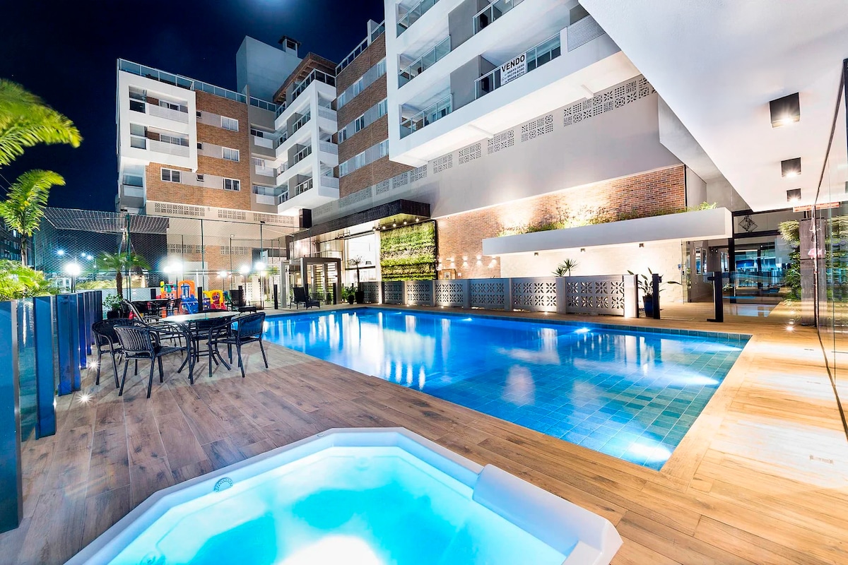 A tranquil outdoor area features a sparkling pool and a heated jacuzzi, surrounded by elegant lighting. Comfortable seating is available nearby, creating an inviting space for relaxation. The modern building architecture complements the serene environment, enhancing the overall appeal of the setting.