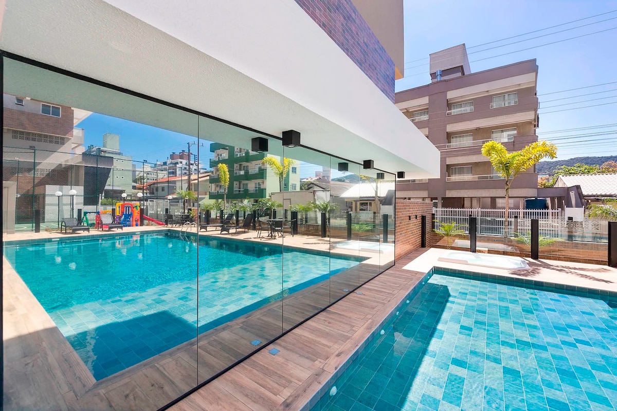 A vibrant outdoor pool area is showcased, featuring clear blue water surrounded by spacious decking. Large glass walls reflect the surrounding scenery. The space includes comfortable lounge chairs arranged neatly and a children's playground visible in the background.