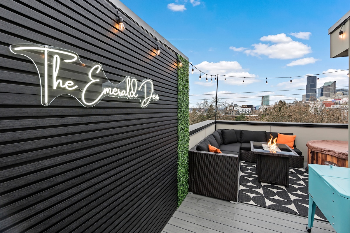 The rooftop deck is furnished with comfortable seating arranged around a propane fire pit. Decorative string lights add ambiance, while the city skyline is visible in the distance. The walls are lined with greenery, enhancing the modern aesthetic of this outdoor space.