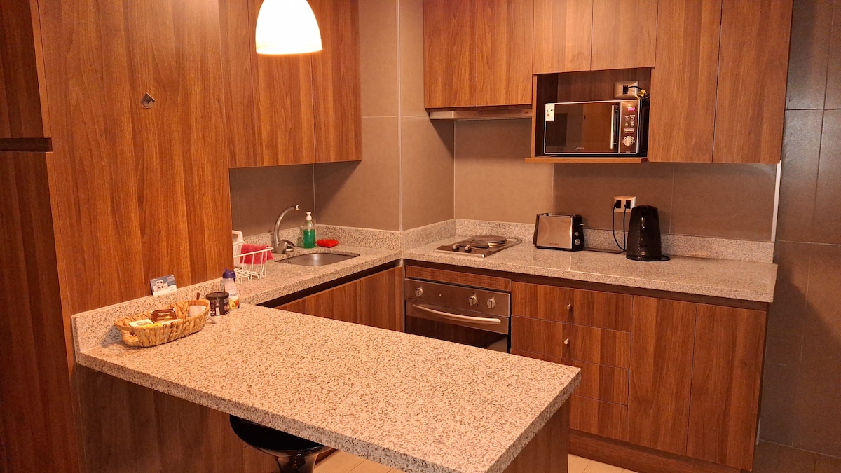A modern kitchen features wooden cabinetry complemented by a granite countertop. Appliances, including a kettle and a microwave, are visible on the countertop. A small breakfast bar provides seating for one person, while a sink and cooking area are neatly organized.