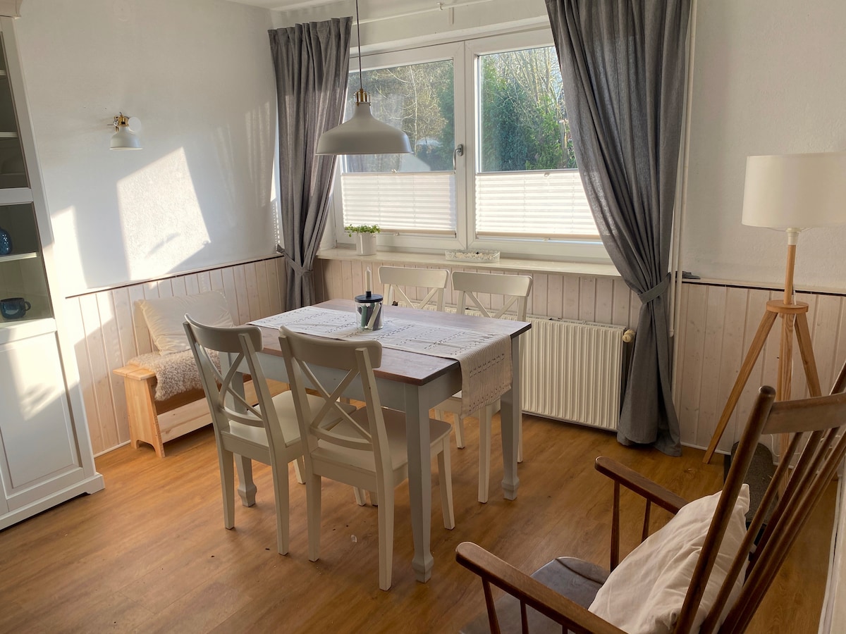 A bright dining area features a wooden table surrounded by four white chairs. Natural light fills the space through a large window covered with light curtains. A cozy armchair and a small side table are positioned nearby, enhancing the room's inviting atmosphere.