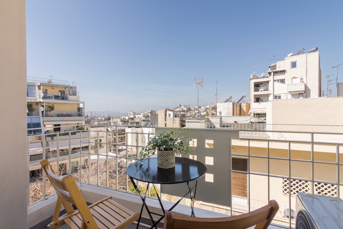 A small balcony area features a round table and two wooden chairs, complemented by a potted plant. The view showcases a mix of urban buildings under a clear blue sky, providing a glimpse of the vibrant neighborhood.