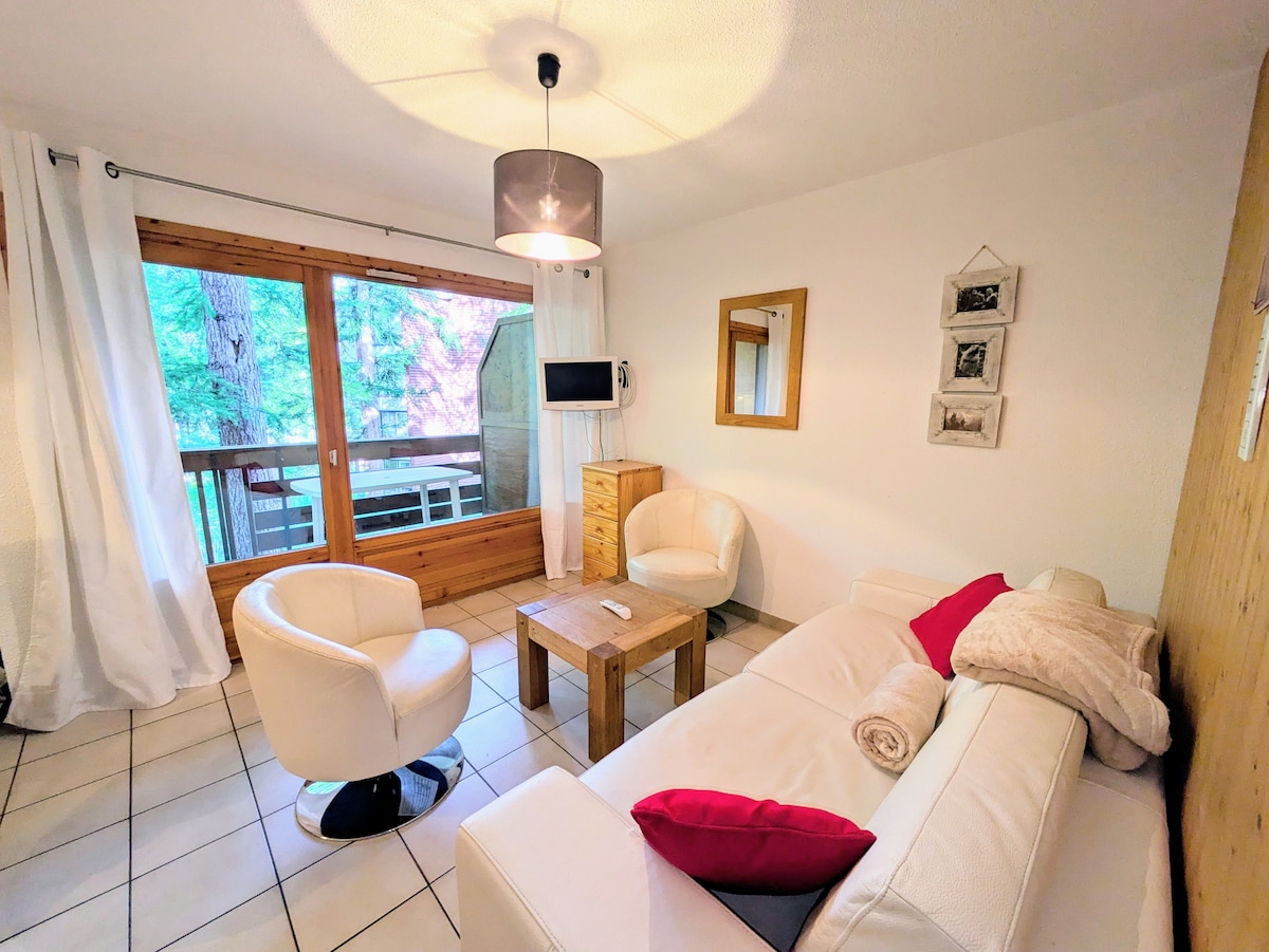 The living area features a cozy white sofa with a textured throw, complemented by two modern armchairs. A wooden coffee table sits at the center, while large windows provide views of the surrounding trees, creating a bright and airy ambiance.