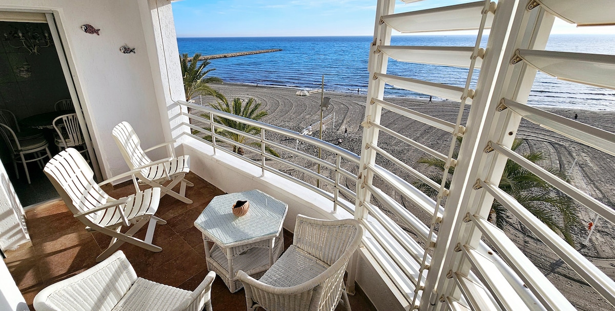 A spacious balcony is furnished with white chairs and a small glass table, offering a panoramic view of the beach and sea. Sunlight reflects off the water, while palm trees are visible nearby, creating a serene outdoor space for relaxation.