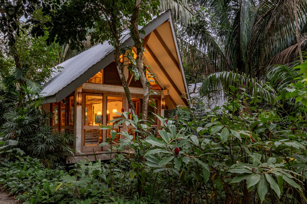 A cozy shala nestled among lush greenery features large glass doors that offer views of the surrounding tropical landscape. The sloped roof adds architectural interest while natural light spills into the inviting interior, enhancing the serene atmosphere of this private retreat.