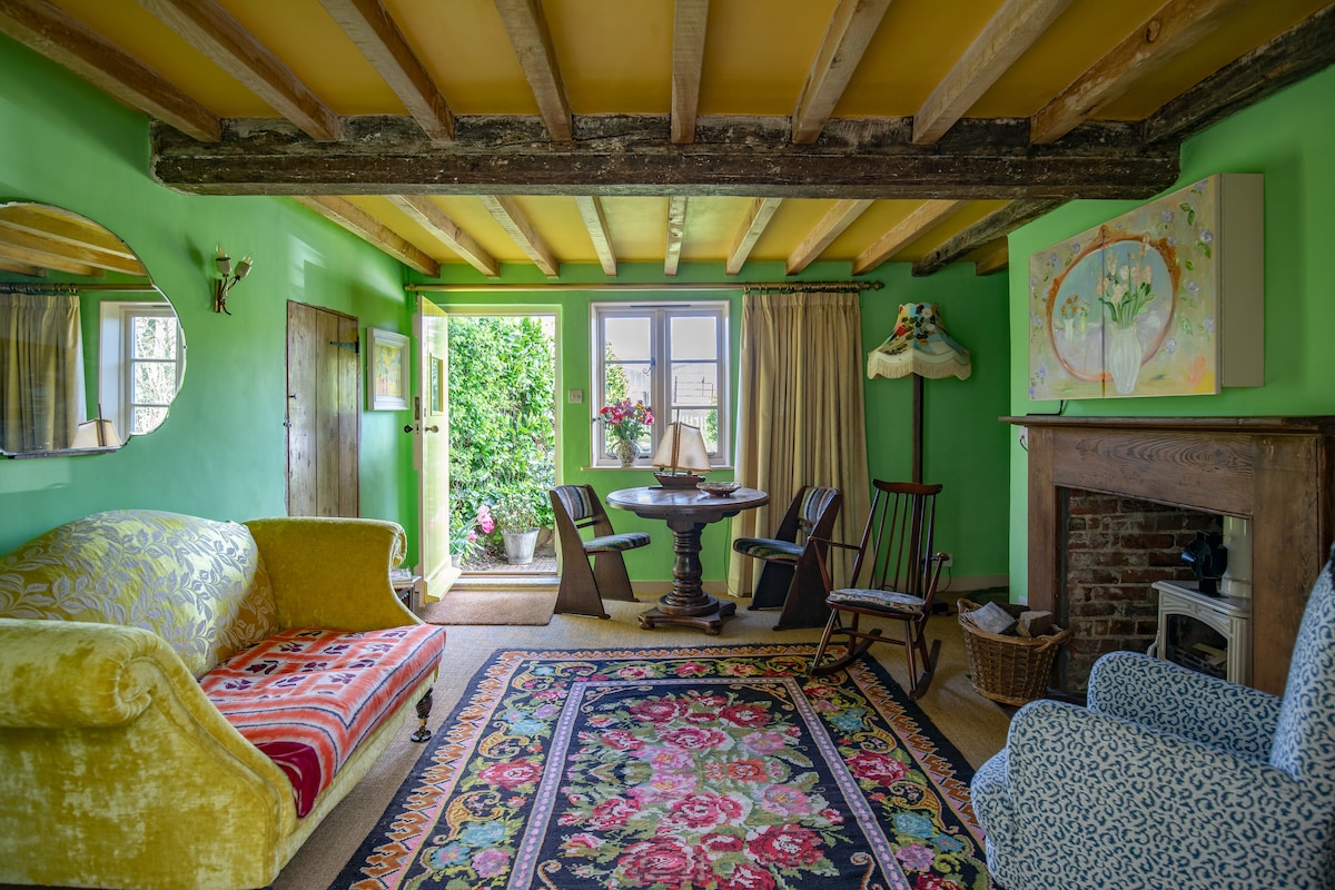 A sitting room features vibrant green walls adorned with original artwork. A cozy yellow sofa and a patterned rug complement the space, alongside a wooden table and chairs positioned by the door. Natural light streams in from the doorway, leading to the garden.
