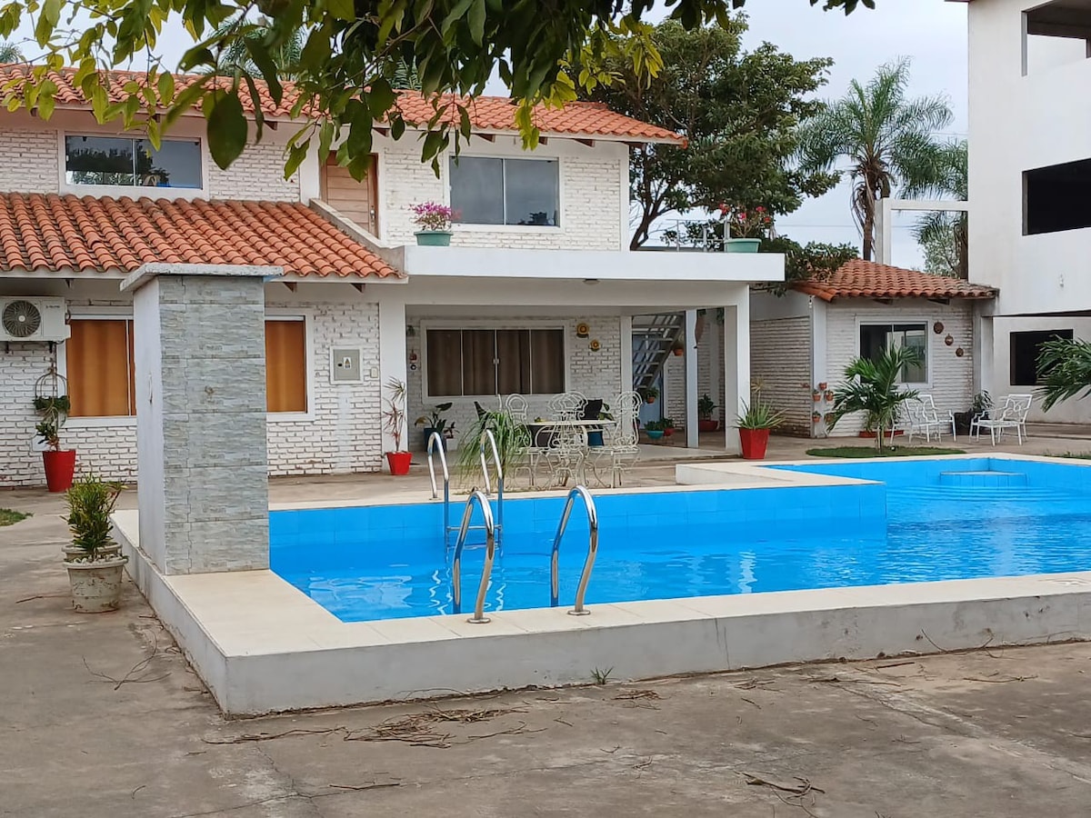 A clear view of a tranquil swimming pool is showcased in the foreground, featuring steps leading into the water. The surrounding area is adorned with potted plants and greenery, while a two-story building stands in the background with a tiled roof, large windows, and a patio area.